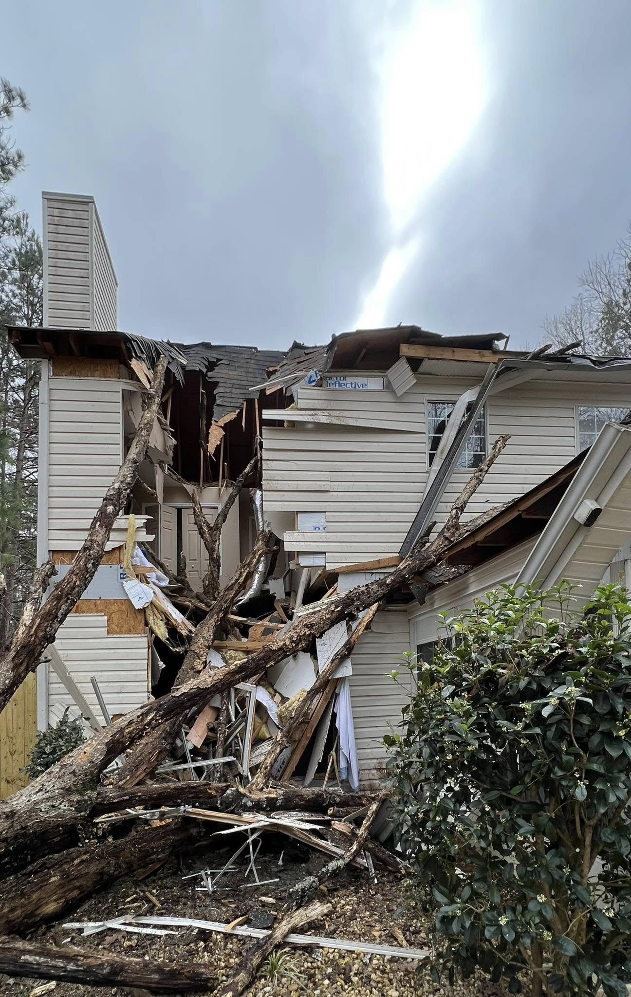 A large tree has fallen onto the roof of a two-story house, causing severe damage and exposing the interior to the outside. Debris is scattered around the scene.