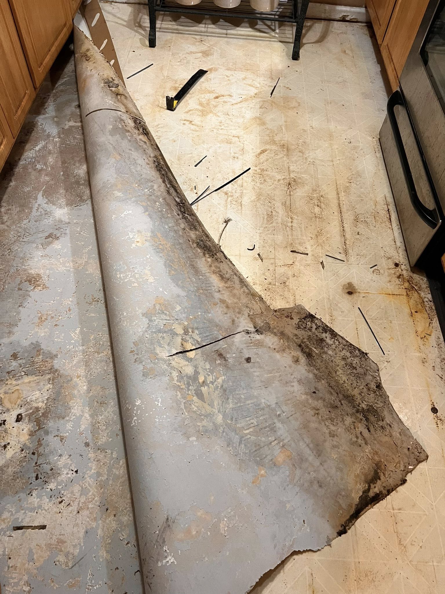 A dirty, stained sheet of old flooring partially peeled back in a kitchen, revealing more stains and dirt on the floor underneath. Tools and debris are scattered around.