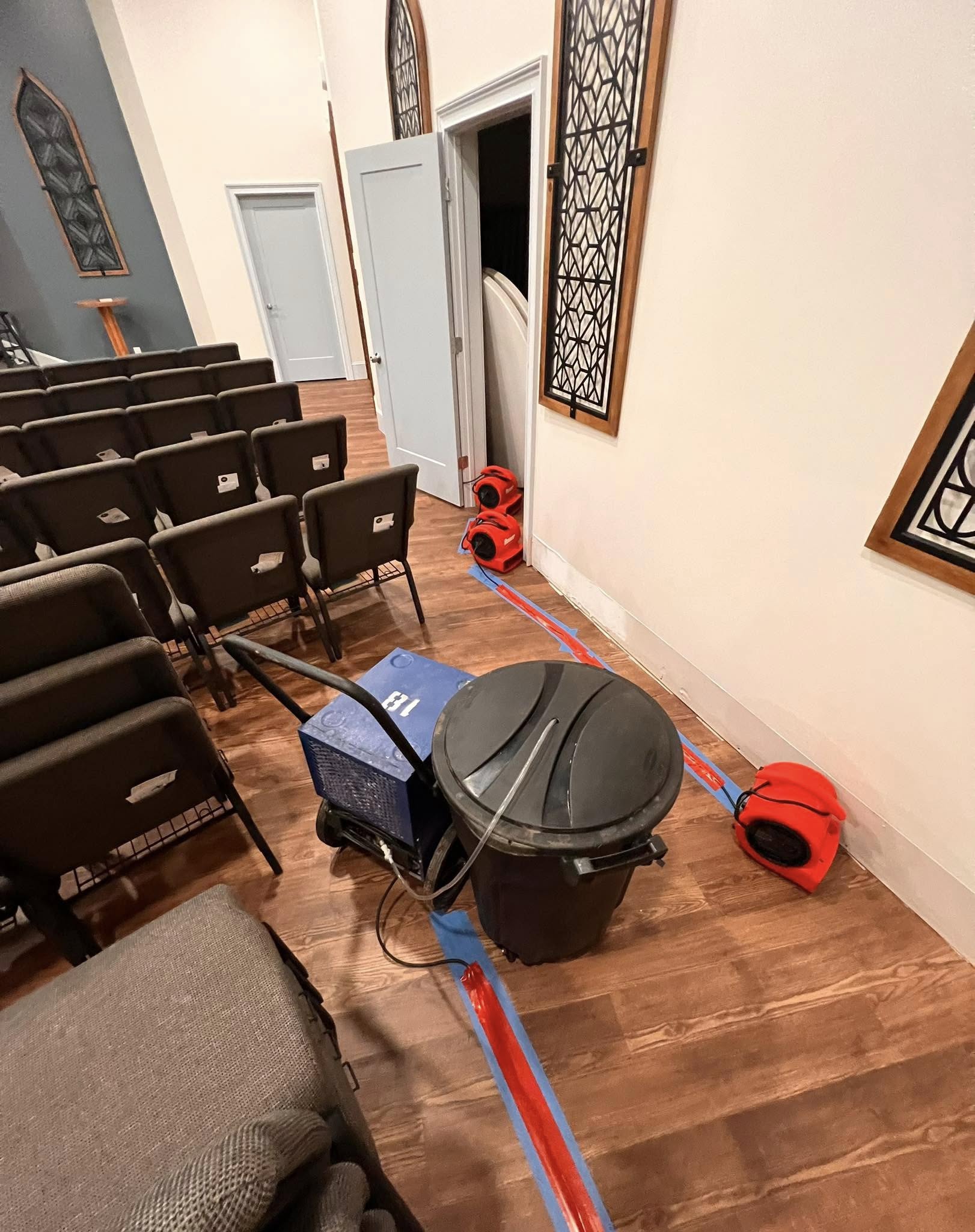 A row of seats in a room with multiple air movers and a dehumidifier on the wooden floor, blue and red tape marking an area near the wall and door.